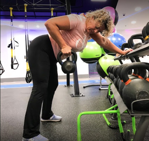 Kobieta w różowej koszulce i czarnych legginsach wykonuje ćwiczenie wiosłowania jednorącz z kettlebellem w siłowni z niebieskim oświetleniem, obok stojaka z kettlebellami i piłkami do ćwiczeń.