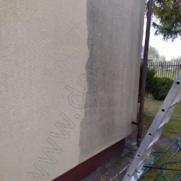 Fragment elewacji budynku z wyraźną różnicą w czystości, gdzie połowa ściany jest umyta, a druga brudna, z widoczną drabiną opartą o ścianę i wężem ogrodowym na ziemi.