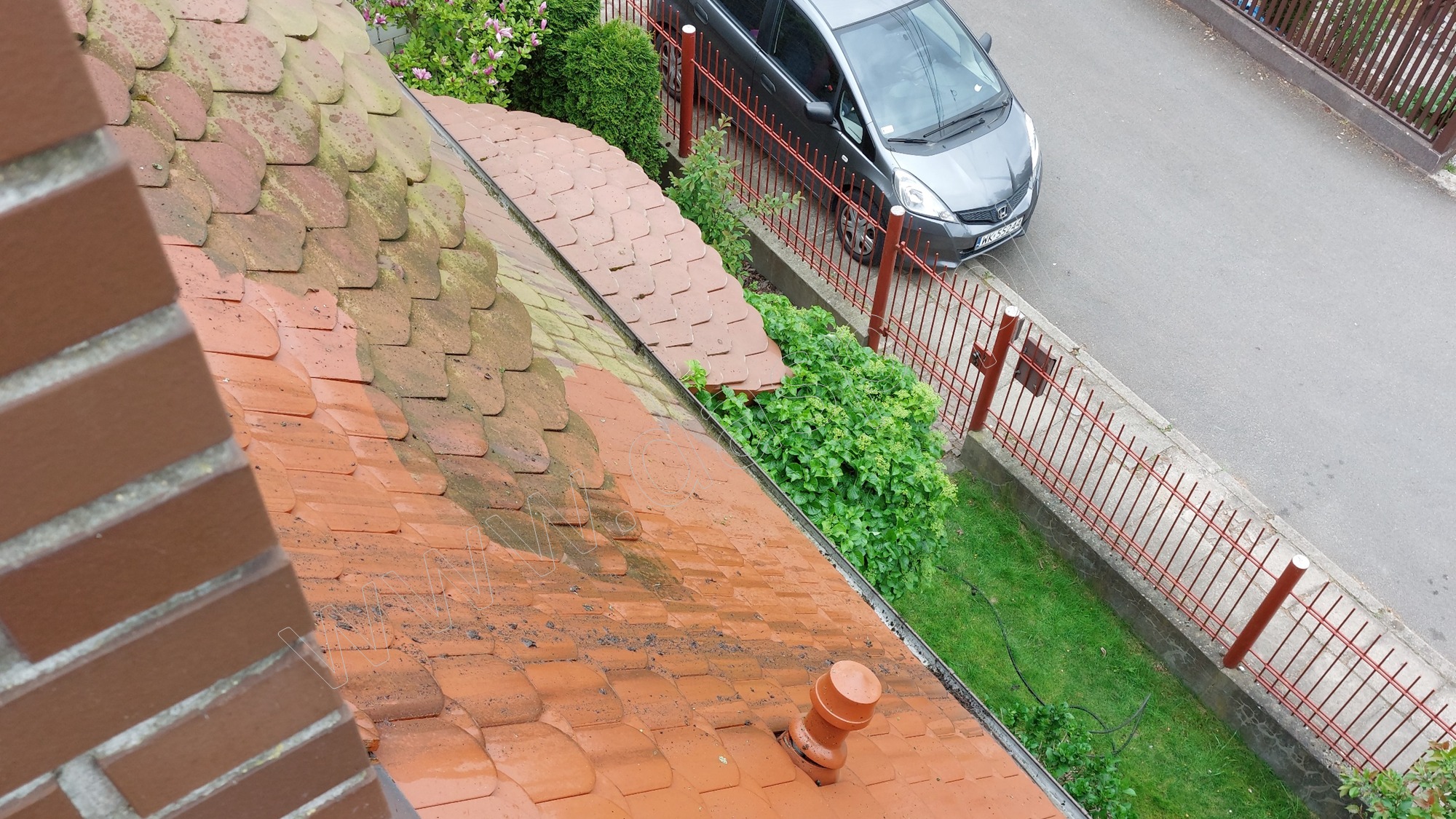 Częściowo oczyszczony dach z czerwonej dachówki, widoczny kontrast między brudną a umytą powierzchnią, w tle szary samochód i ogrodzenie.