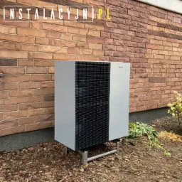 Zewnętrzna jednostka pompy ciepła Viessmann zamontowana przy ceglanej ścianie budynku, widoczne metalowe nóżki i ściółka z kory.