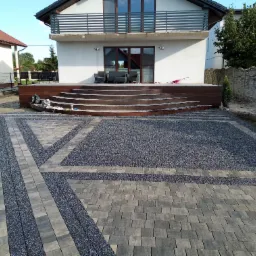 Nowoczesny podjazd z szarej kostki brukowej ułożonej we wzór, z drewnianym tarasem i schodami prowadzącymi do domu z balkonem.