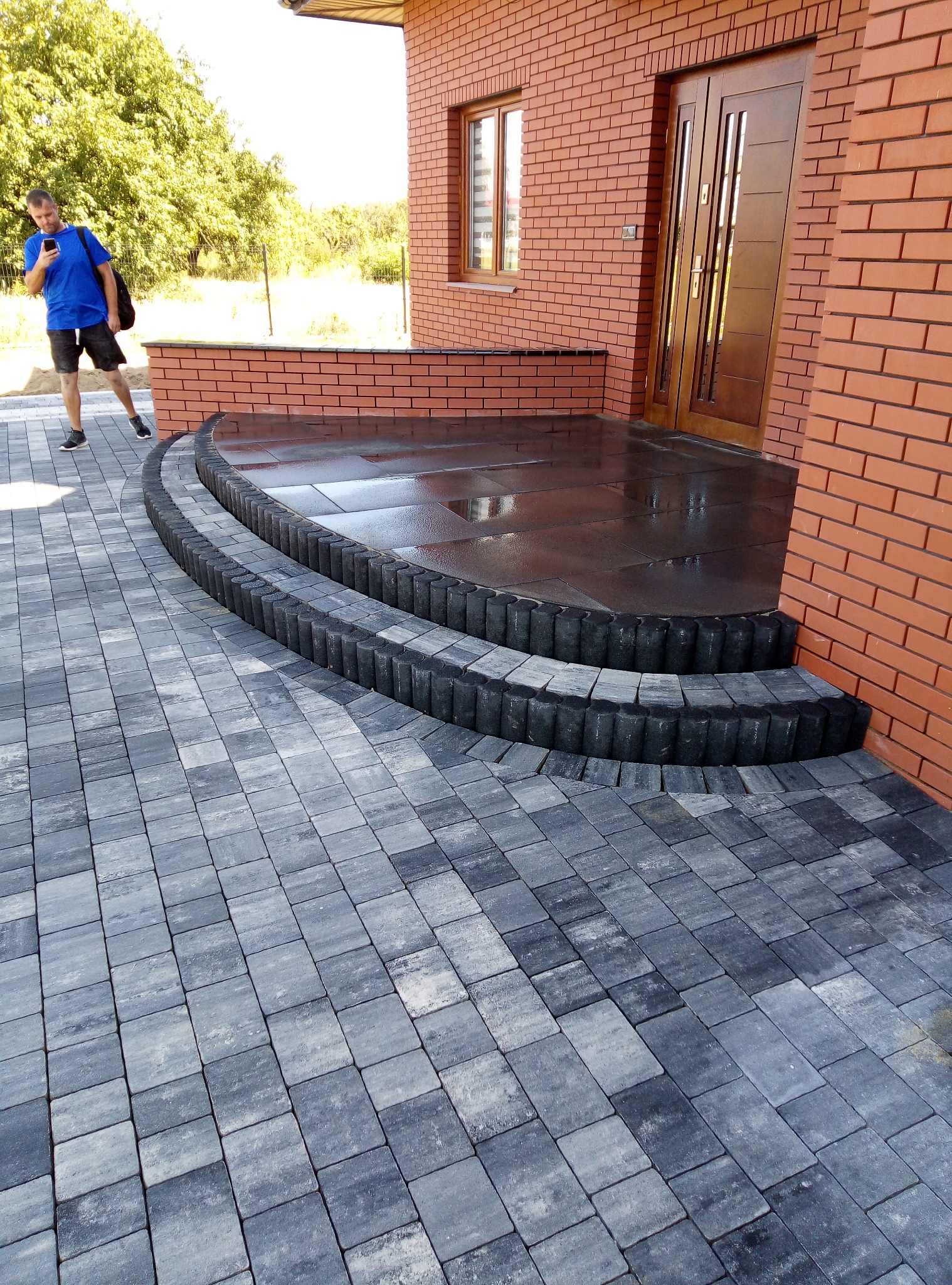 Ułożona szara kostka brukowa prowadząca do dwustopniowych schodów z ciemnych obrzeży, mokry taras przed budynkiem z cegły, mężczyzna z telefonem w tle.