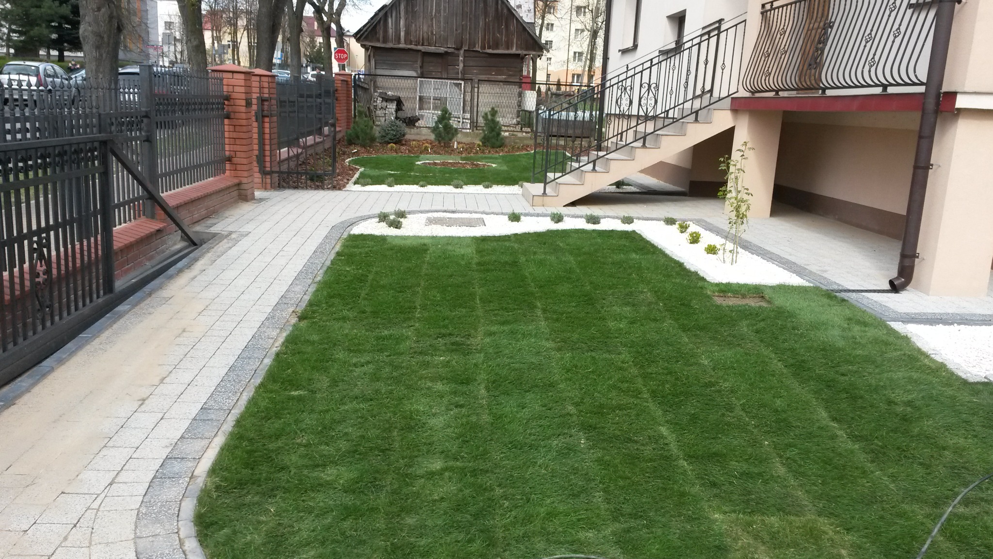 Nowo założony trawnik przed domem z kostką brukową na obrzeżach i dekoracyjnym białym kamieniem, widoczne schody zewnętrzne i ogrodzenie z cegły.