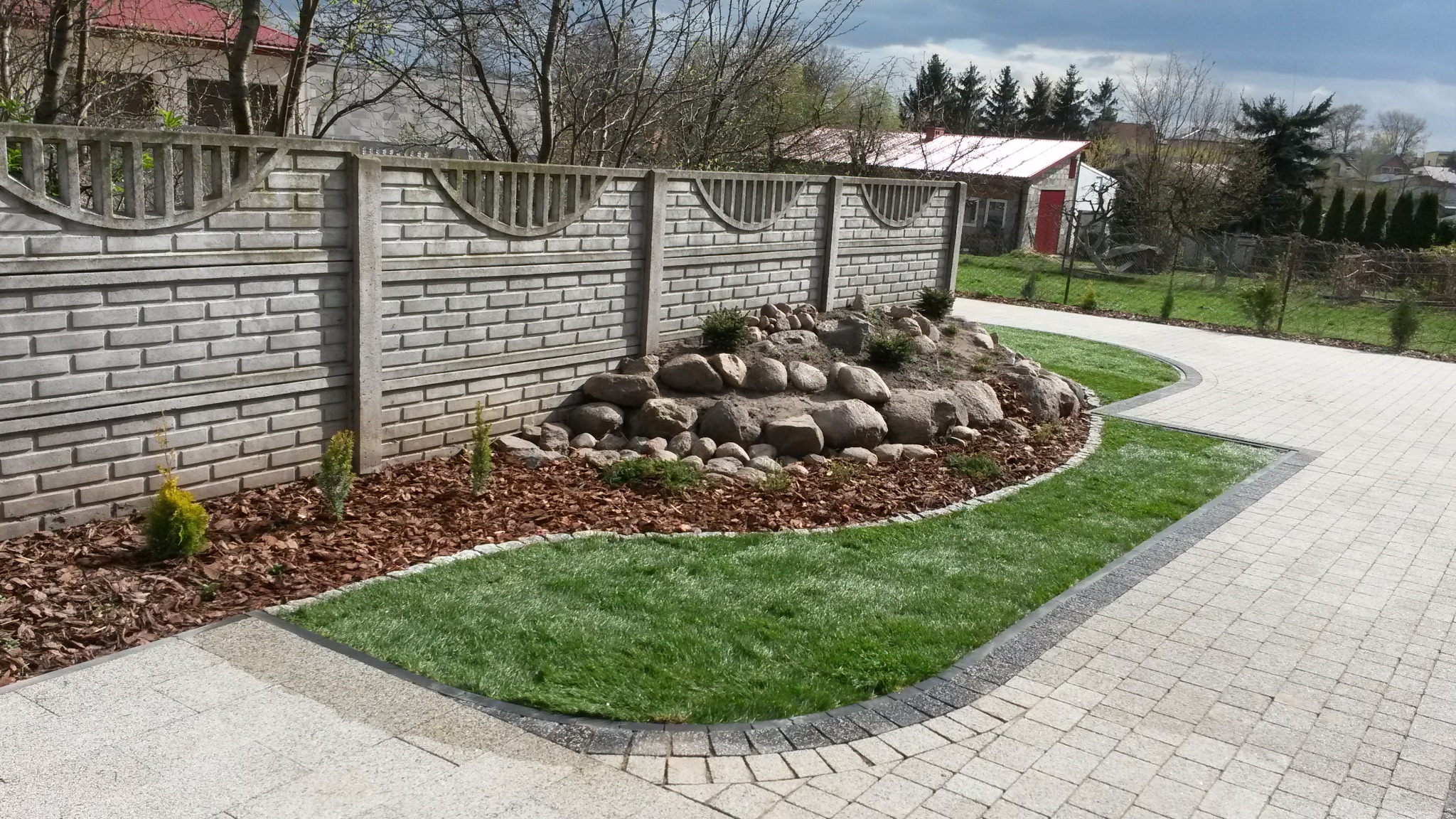 Ścieżka z jasnej kostki brukowej obramowana ciemniejszymi kostkami, przylegająca do trawnika z rabatą z kamieni i kory, w tle betonowy płot z ozdobnymi wycięciami.