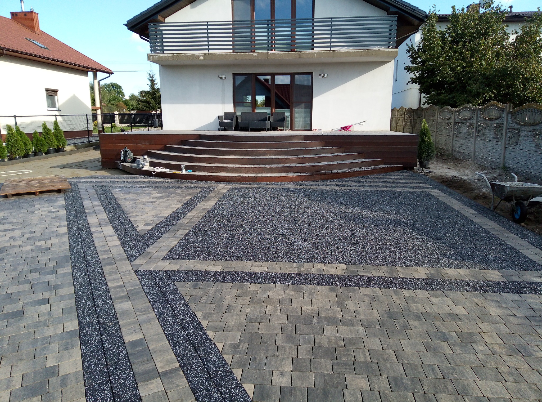 Nowoczesny podjazd z szarej kostki brukowej ułożonej we wzór, z drewnianym tarasem i schodami prowadzącymi do domu z balkonem.