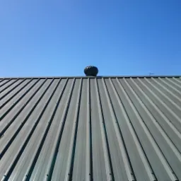 Szary dach z blachy trapezowej z turbiną wiatrową na tle błękitnego nieba.