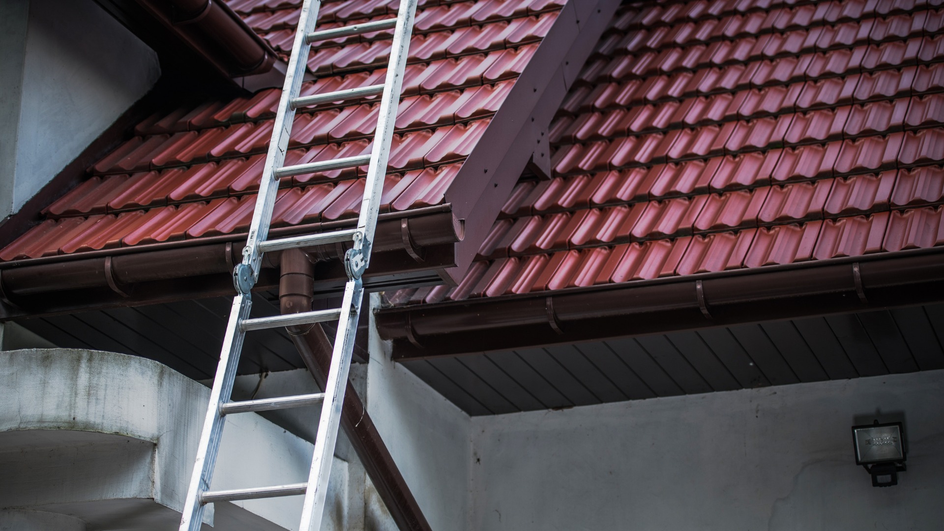 Aluminiowa drabina oparta o brązową rynnę i dach pokryty czerwoną dachówką ceramiczną.