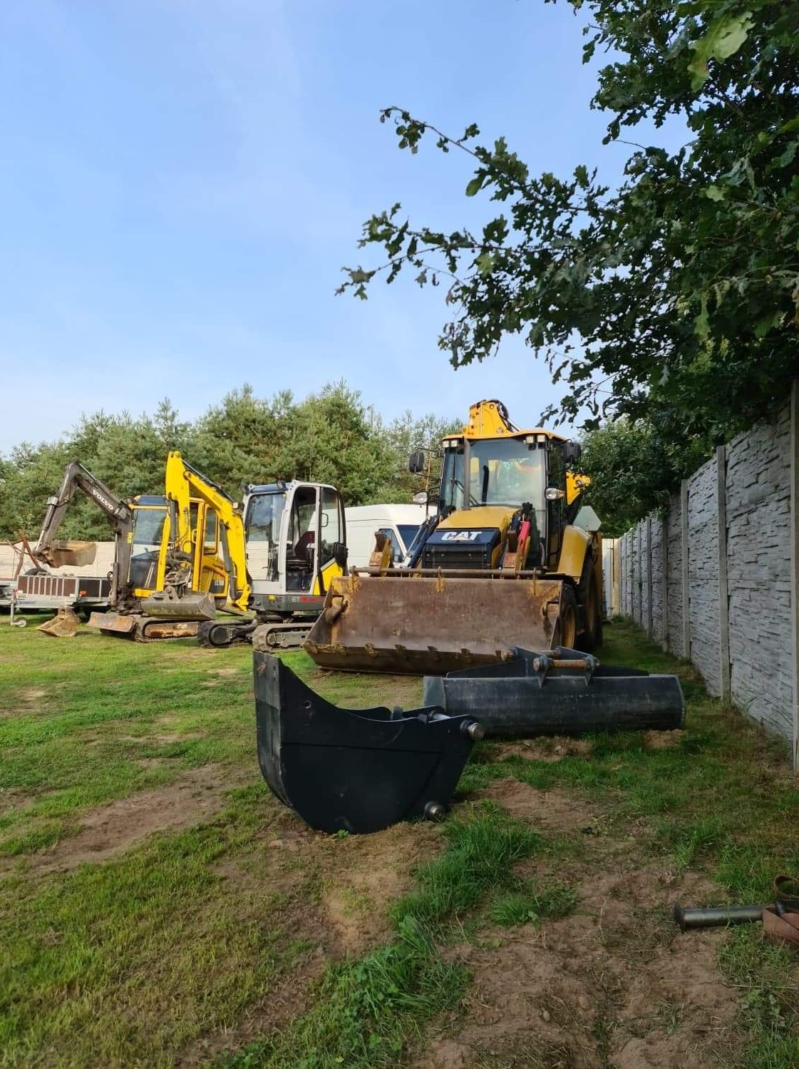 Żółta koparko-ładowarka CAT i dwie mniejsze koparki na trawiastym terenie obok betonowego ogrodzenia panelowego, dodatkowe łyżki koparkowe leżą na ziemi.