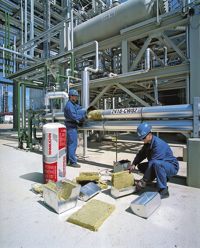 Pracownicy w niebieskich kombinezonach izolują rury przemysłowe wełną mineralną Rockwool, widoczne metalowe korytka i rolka materiału izolacyjnego, w tle skomplikowana konstrukcja rurociągów.