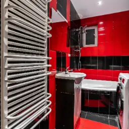 Nowoczesna łazienka z czerwono-czarnymi płytkami, białym grzejnikiem drabinkowym, umywalką z szafką, wanną i pralką.