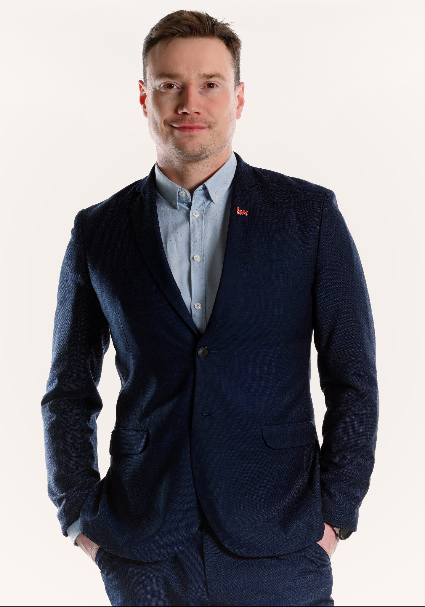 Portret uśmiechniętego mężczyzny w granatowym garniturze i błękitnej koszuli, z rękami w kieszeniach, prezentującego się profesjonalnie z przypinką 'kw' na klapie.