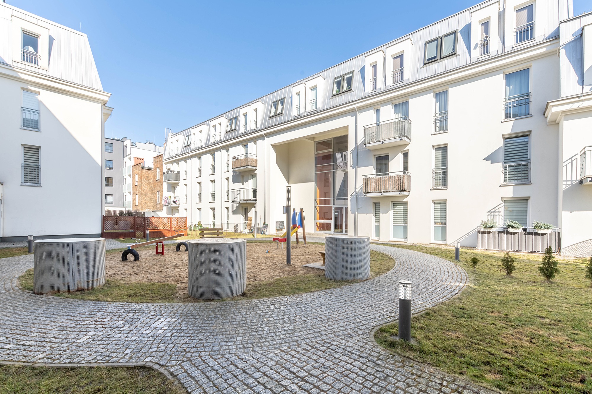 Nowoczesny kompleks mieszkalny z wewnętrznym dziedzińcem i placem zabaw, widok na budynki z balkonami, brukowaną ścieżkę i piaskownicę z huśtawką.