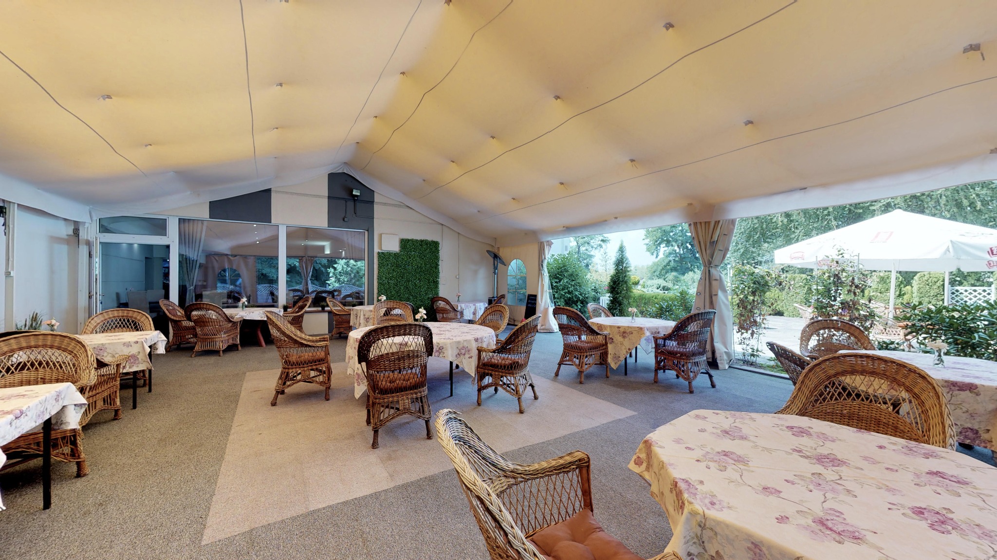 Zadaszony taras z wiklinowymi krzesłami i stolikami nakrytymi kwiecistymi obrusami, widok na ogród z białym parasolem w tle.