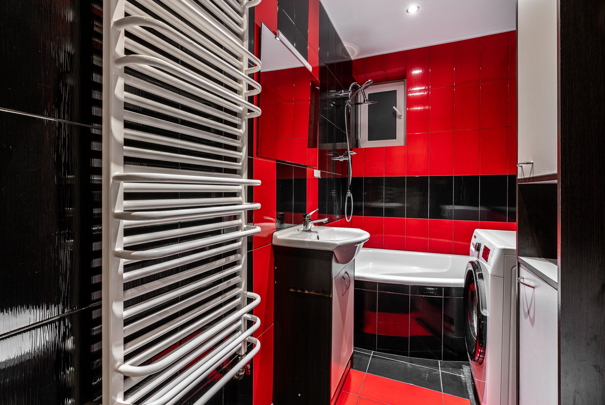 Nowoczesna łazienka z czerwono-czarnymi płytkami, białym grzejnikiem drabinkowym, umywalką z szafką, wanną i pralką.