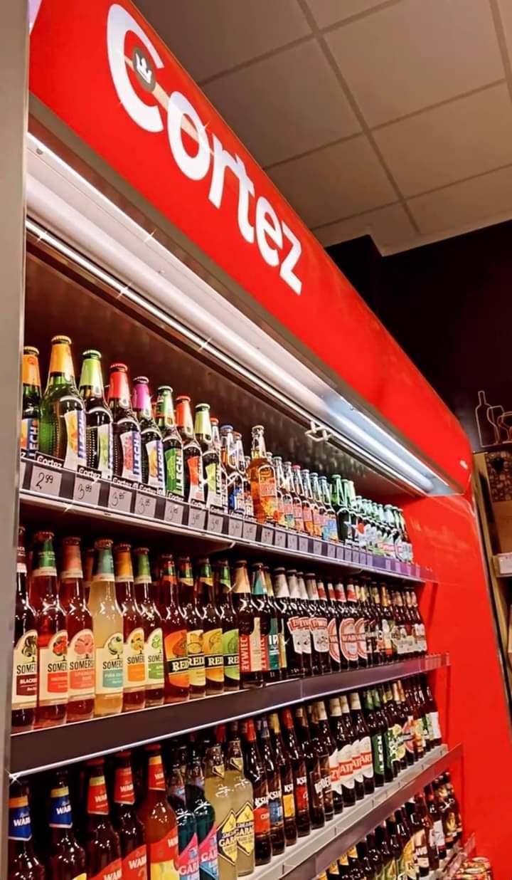 Półki chłodnicze wypełnione różnorodnymi butelkami piwa i napojów alkoholowych, z widocznym logo 'Cortez' na czerwonym panelu nad nimi.