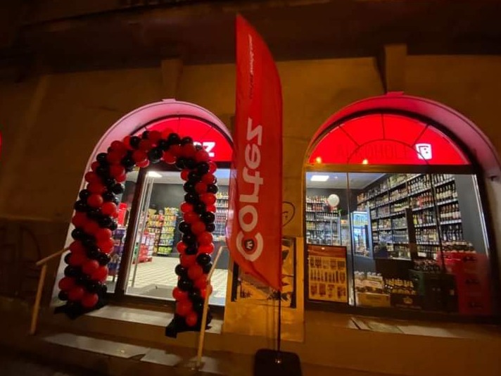 Wejście do sklepu z alkoholem udekorowane łukiem z czerwono-czarnych balonów i czerwoną flagą reklamową, widoczne półki pełne butelek wewnątrz.