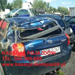 Zniszczony, niebieski samochód Toyota Corolla z uszkodzonym tyłem, widoczne inne wraki aut w tle, na karoserii tekst reklamowy firmy zajmującej się kasacją pojazdów.