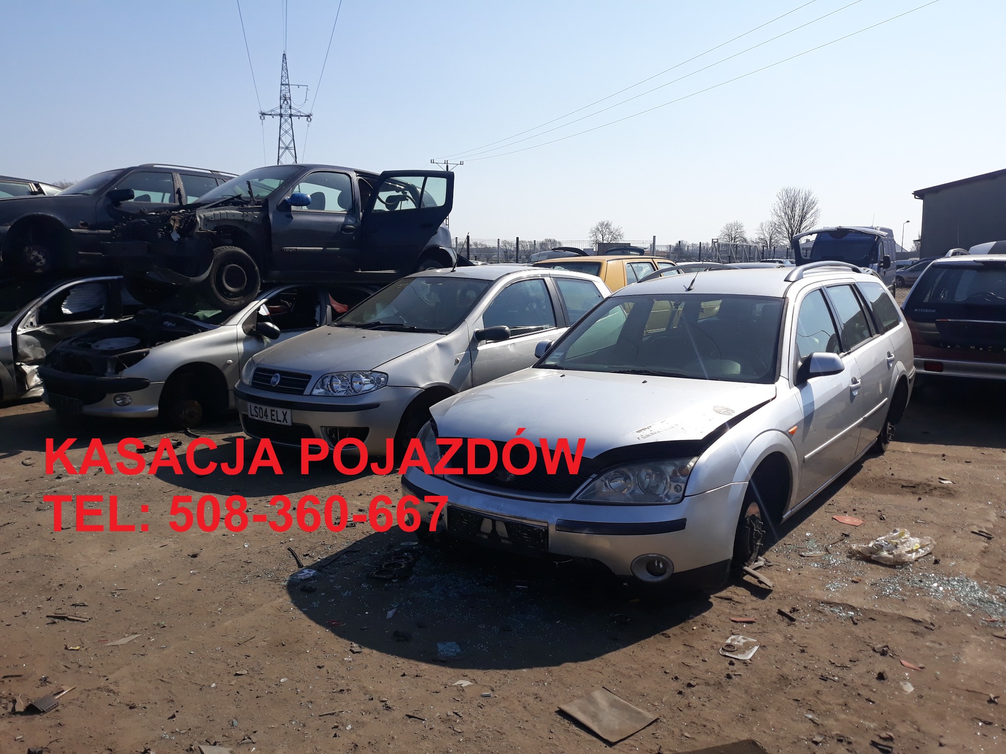 Srebrny Ford Mondeo kombi z uszkodzonym przodem na placu pełnym wraków samochodów, z widoczną linią wysokiego napięcia w tle.