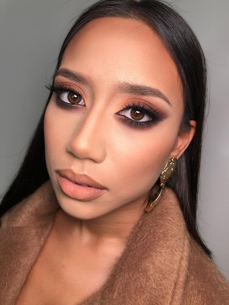 Portret kobiety z wyrazistym, wieczorowym eye-art w odcieniach brązu i beżu, podkreślonymi brwiami i błyszczącą skórą, w beżowym płaszczu i z dużą, złotą kolczykiem.