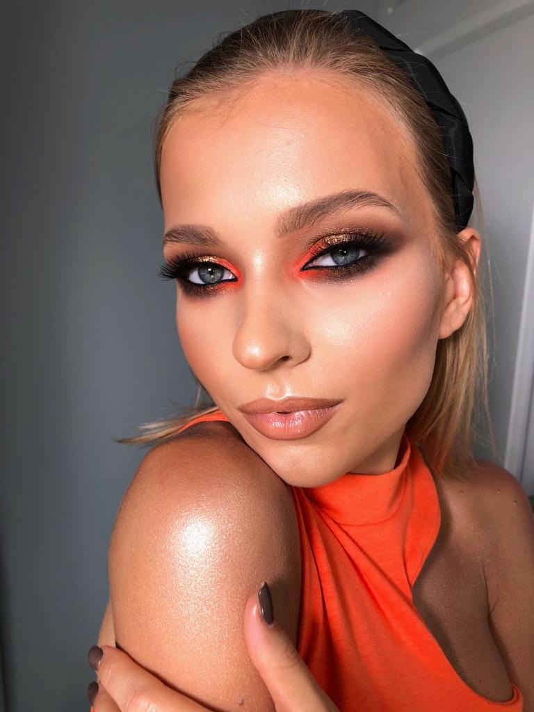 Portret kobiety z intensywnym, wieczorowym w tonacji pomarańczowo-brązowej, błyszczące cienie na powiekach i podkreślone usta błyszczykiem, czarna opaska na włosach.