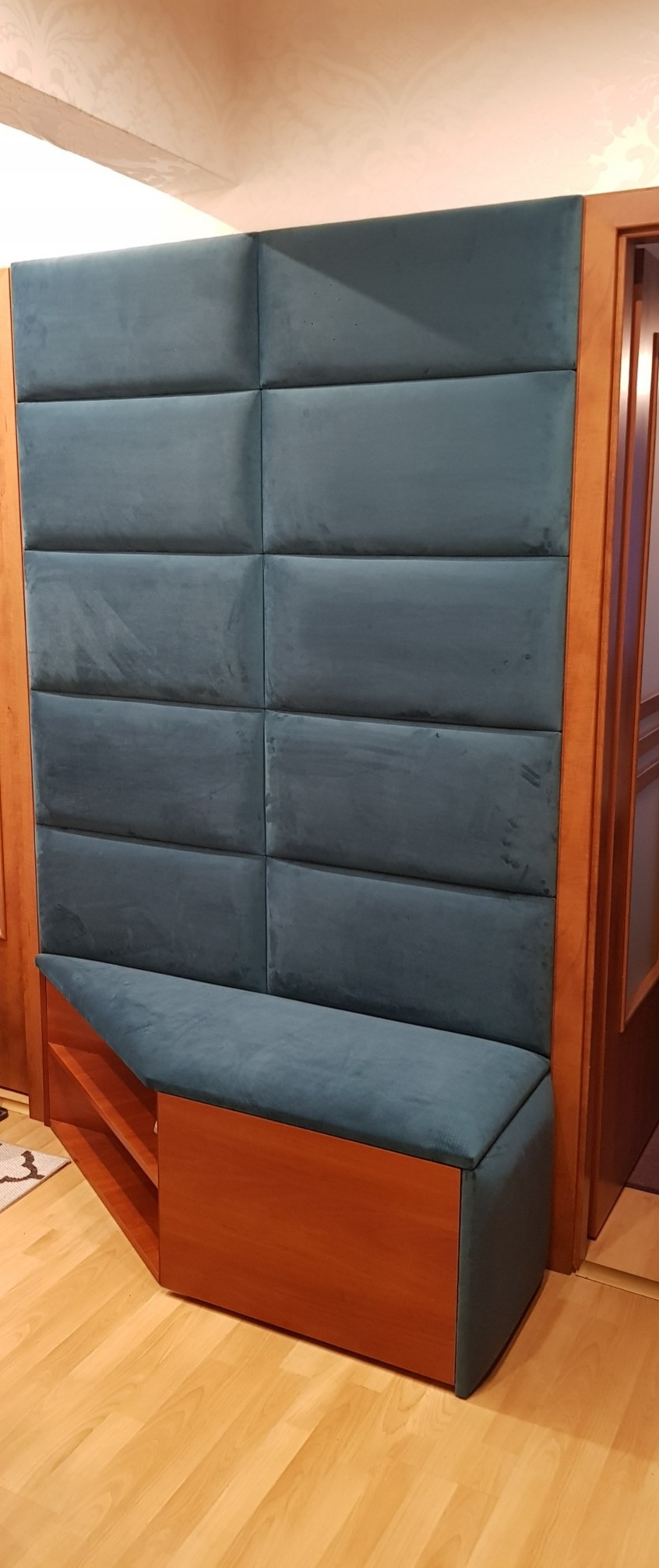 Panel tapicerowany z siedziskiem w kolorze morskim, połączony z drewnianą szafką z półkami, tworzący zabudowę w przedpokoju.