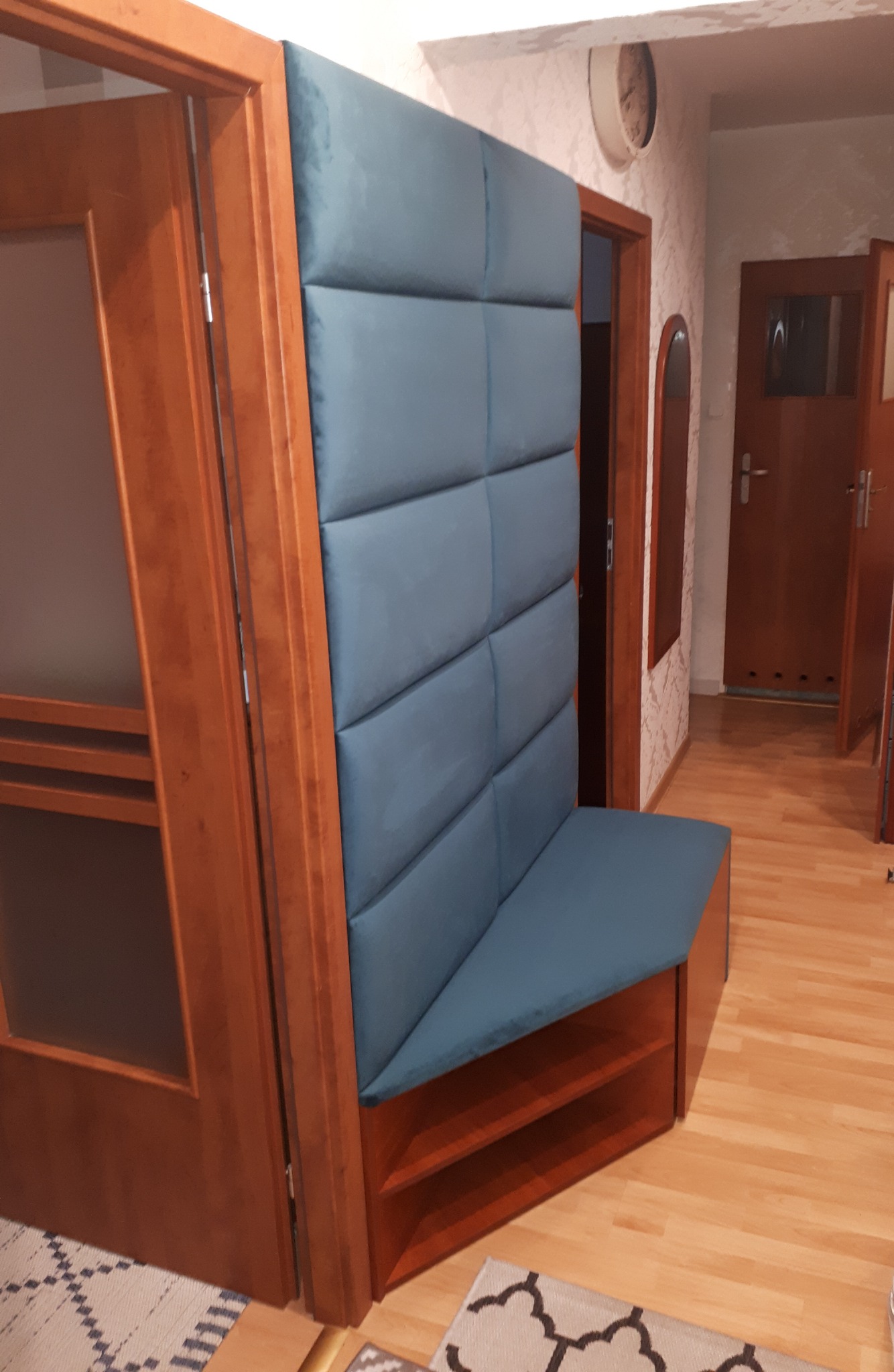 Narożna zabudowa tapicerowana w przedpokoju, z siedziskiem i pionowym panelem w kolorze gołębiej szarości, wkomponowana w drewnianą konstrukcję z półkami.