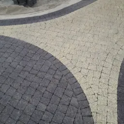Ułożona kostka brukowa w dwóch kolorach, tworząca wzór łuków na powierzchni. Widoczne zapasy piasku i kostki w tle.
