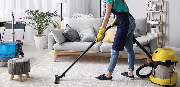 Osoba w niebieskim fartuchu i żółtych rękawicach odkurza jasny dywan w salonie za pomocą żółtego odkurzacza. W tle widoczna sofa, roślina doniczkowa i wózek z akcesoriami do sprzątania.