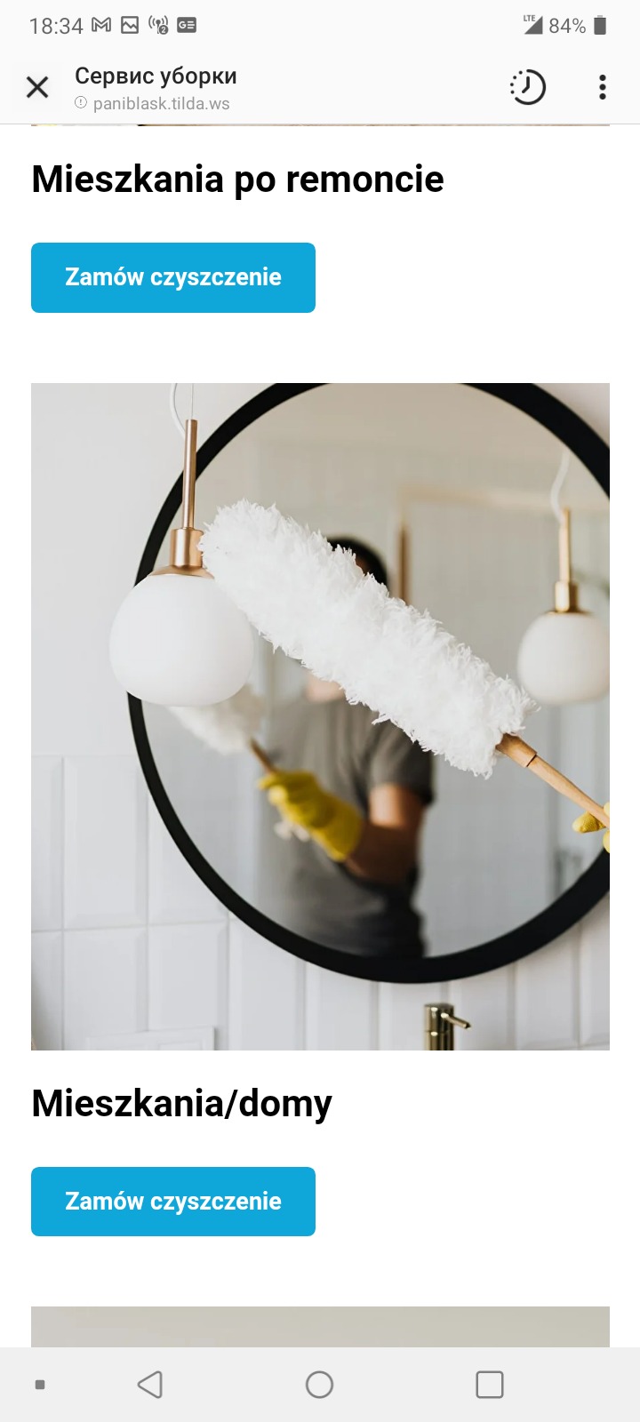 Osoba w żółtych rękawicach czyści okrągłe lustro za pomocą miotełki z piór, widoczne białe kafelki na ścianie i złota armatura.