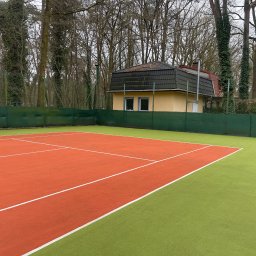 CDM COMPANY SP. Z O.O. - Czerwono-zielony kort tenisowy z widocznym budynkiem w tle, otoczony siatką i drzewami. Widok na czystą nawierzchnię kortu po renowacji.