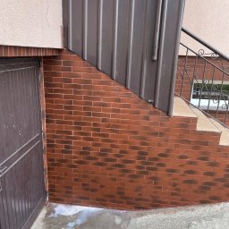 CDM COMPANY SP. Z O.O. - Fragment elewacji z brązowej cegły klinkierowej, schody z metalową barierką, blaszane obicie i ślady po czyszczeniu na betonowej posadzce przy kratce ściekowej.