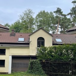 CDM COMPANY SP. Z O.O. - Jasna elewacja domu z brązowym dachem i oknami, widoczna zza metalowego ogrodzenia porośniętego bluszczem. Na dachu okna połaciowe i komin.