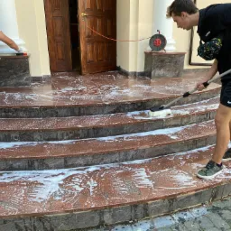 Mężczyzna czyści schody wejściowe z czerwonej kostki brukowej za pomocą szczotki na kiju, drugi mężczyzna czyści kolumnę obok drzwi wejściowych, widoczna piana po czyszczeniu i zwinięty przedłużacz.