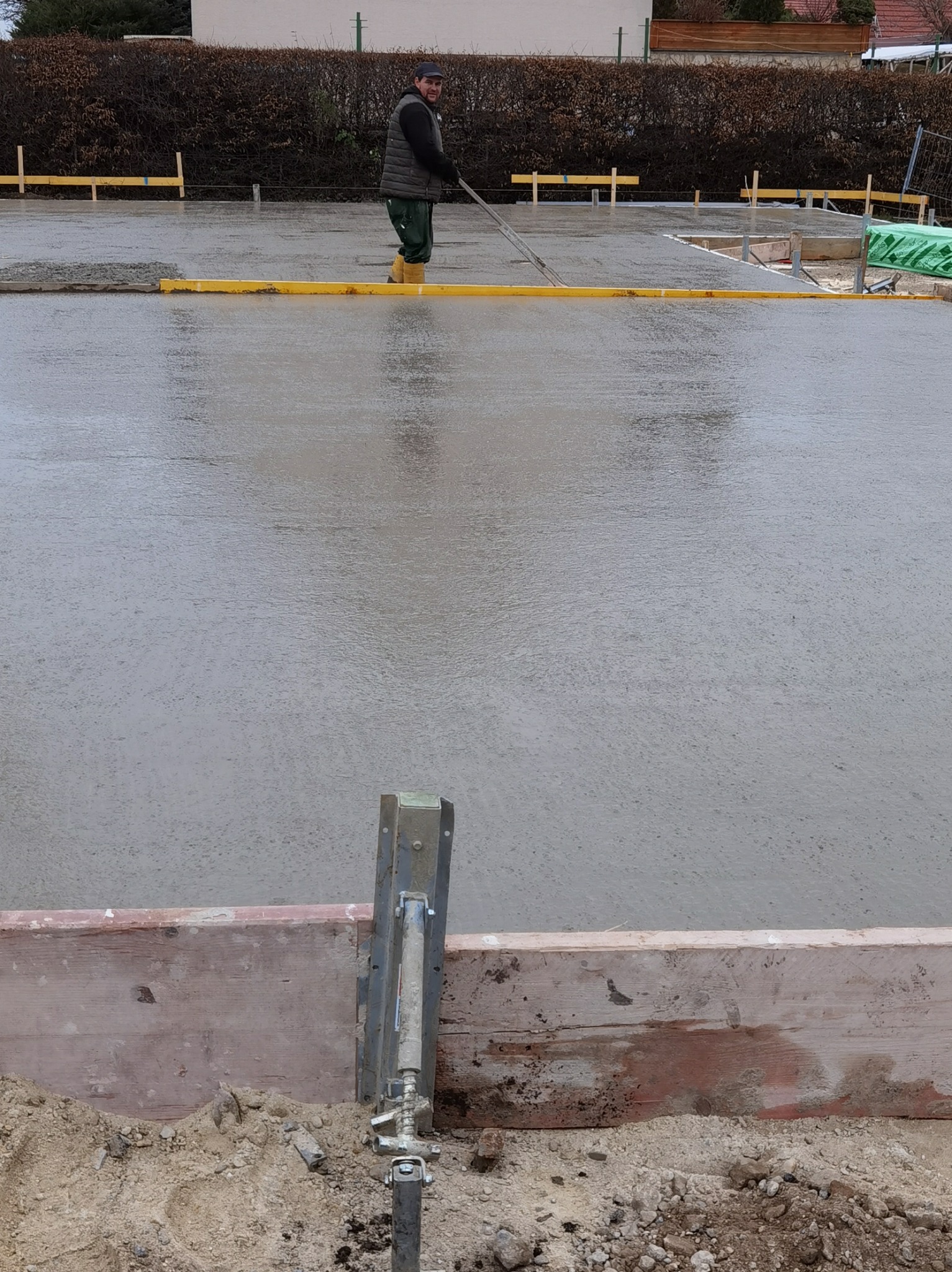 Pracownik w kaloszach wyrównuje wylewkę betonową za pomocą długiej listwy, widoczne drewniane szalunki i poziomice.