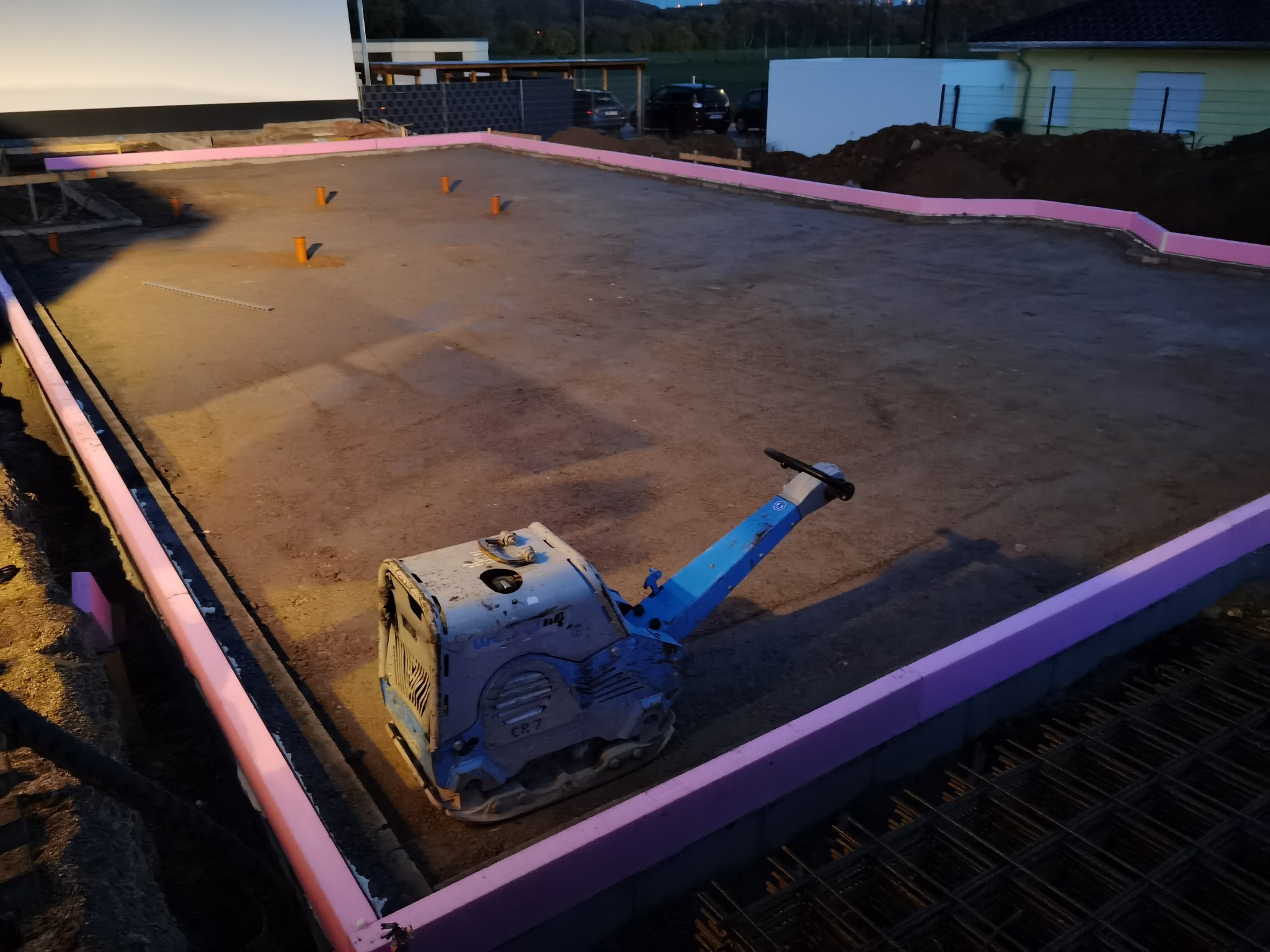 Przygotowanie podłoża pod fundament: ubijarka wibrująca na wyrównanym terenie, obrzeża z różowego styropianu, wystające rury i siatka zbrojeniowa.