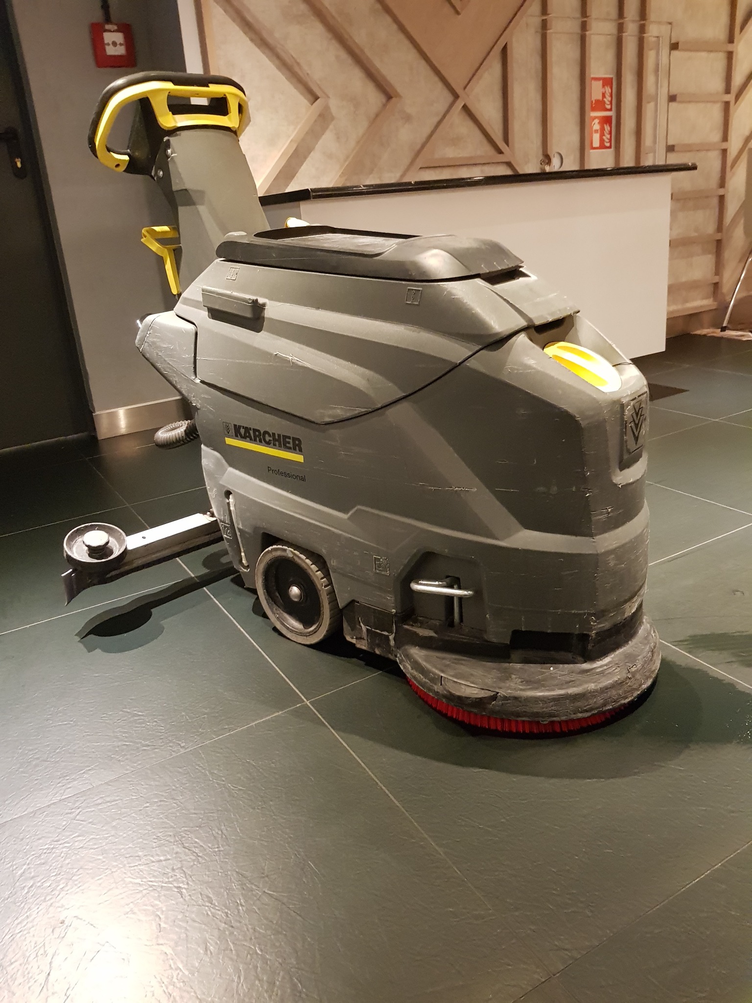 Profesjonalna szorowarka Kärcher z żółtymi akcentami, stojąca na ciemnej, błyszczącej posadzce w pomieszczeniu z elementami dekoracyjnymi na ścianie.