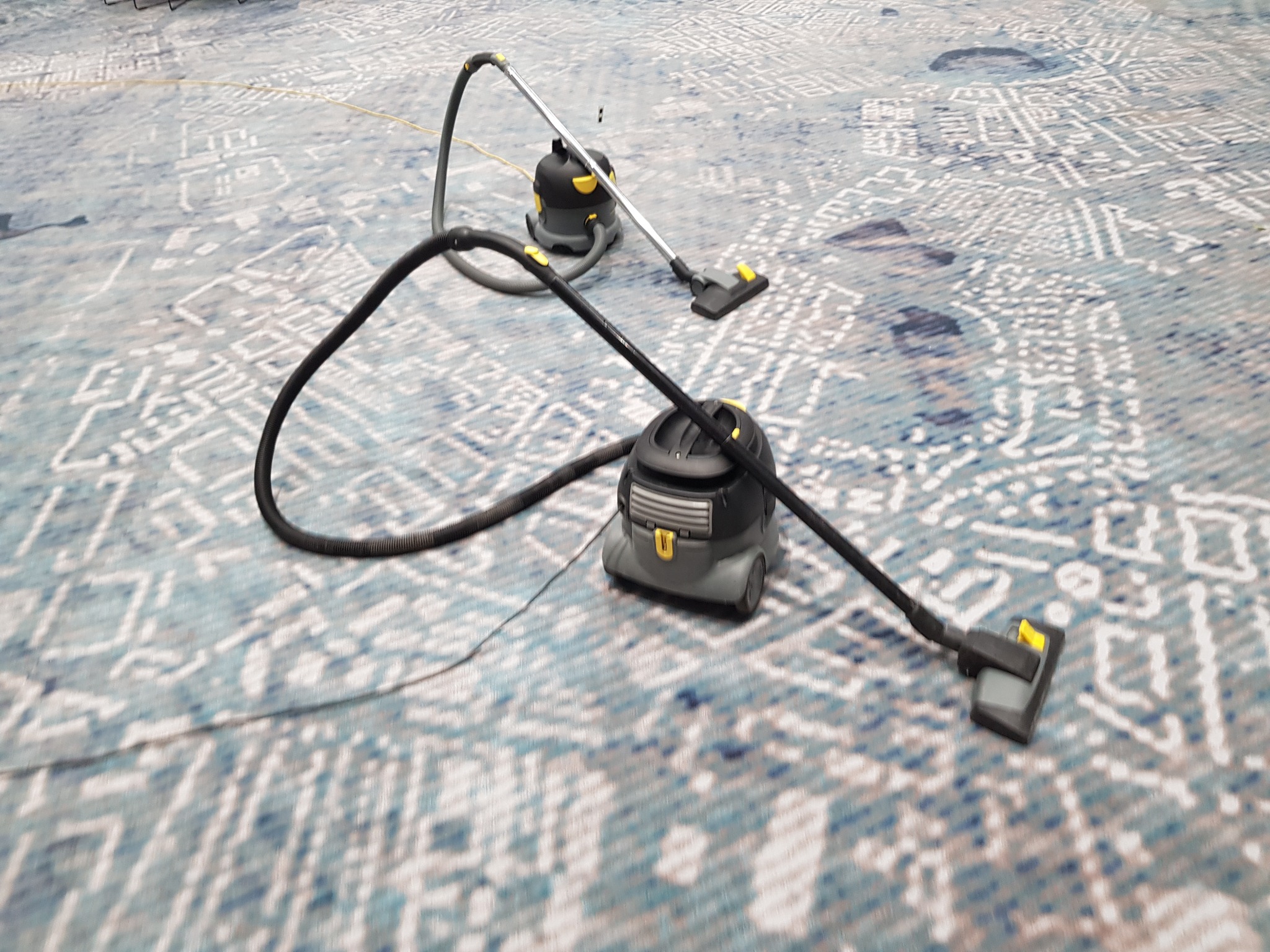 Dwa odkurzacze piorące na wzorzystym dywanie, jeden z wężem na pierwszym planie, drugi z rurą w tle.