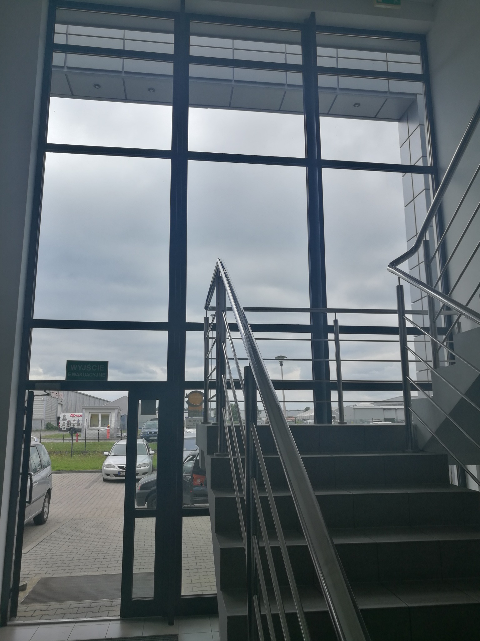 Klatka schodowa z metalową balustradą i dużym oknem z widokiem na parking i pochmurne niebo.