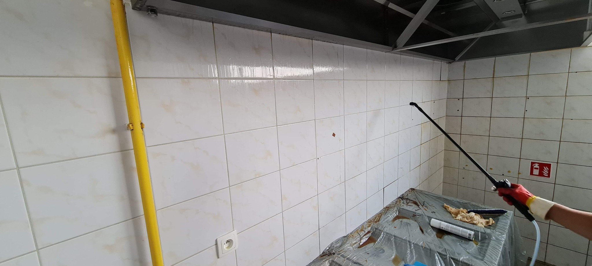 Czyszczenie płytek ściennych w pomieszczeniu roboczym za pomocą lancy spryskującej, widoczny fragment żółtej rury gazowej i osprzęt pod sufitem.