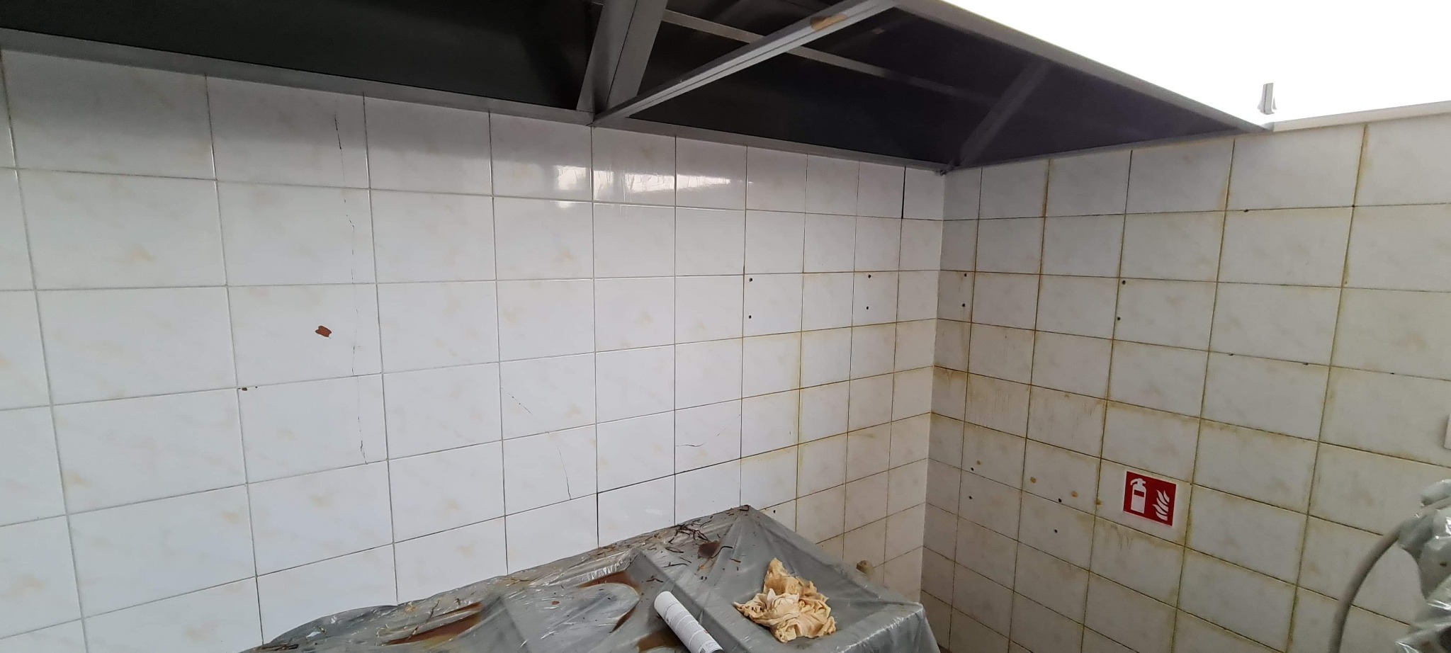 Brudne, białe kafelki z brązowymi zaciekami w rogu pomieszczenia, zabezpieczone folią meble i widoczny znak gaśnicy na ścianie.
