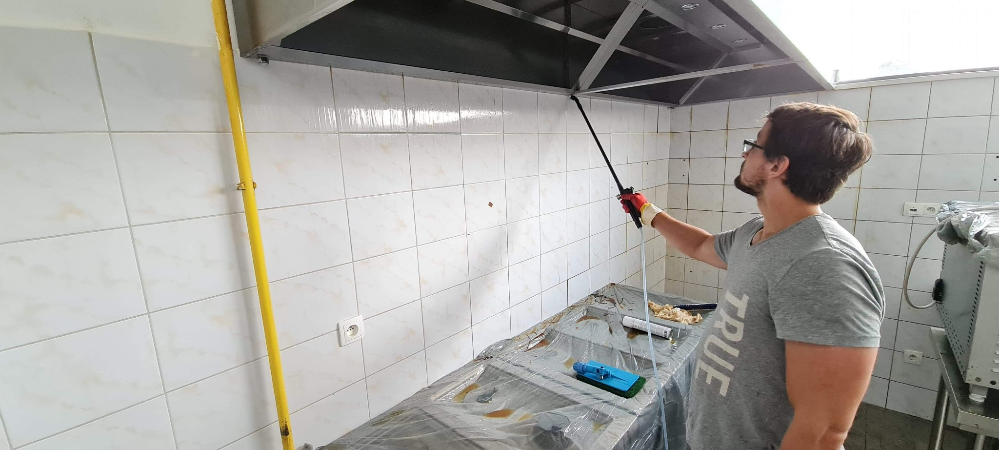 Mężczyzna w okularach czyści okap kuchenny w lokalu gastronomicznym za pomocą lancy i środka czyszczącego, zabezpieczone blaty folią ochronną.