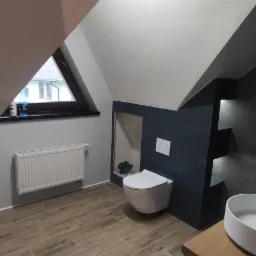 Nowoczesna łazienka na poddaszu z drewnopodobną podłogą, wiszącą toaletą i umywalką nablatową, wykończona ciemnymi płytkami z wnękami.