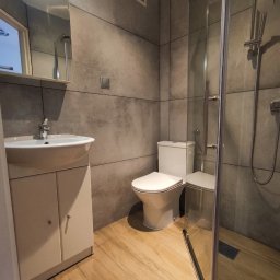 ANATOLII - Nowoczesna łazienka z kabiną prysznicową, umywalką z szafką i toaletą. Szare płytki imitujące beton na ścianach, drewnopodobne panele na podłodze. Minimalistyczny design.