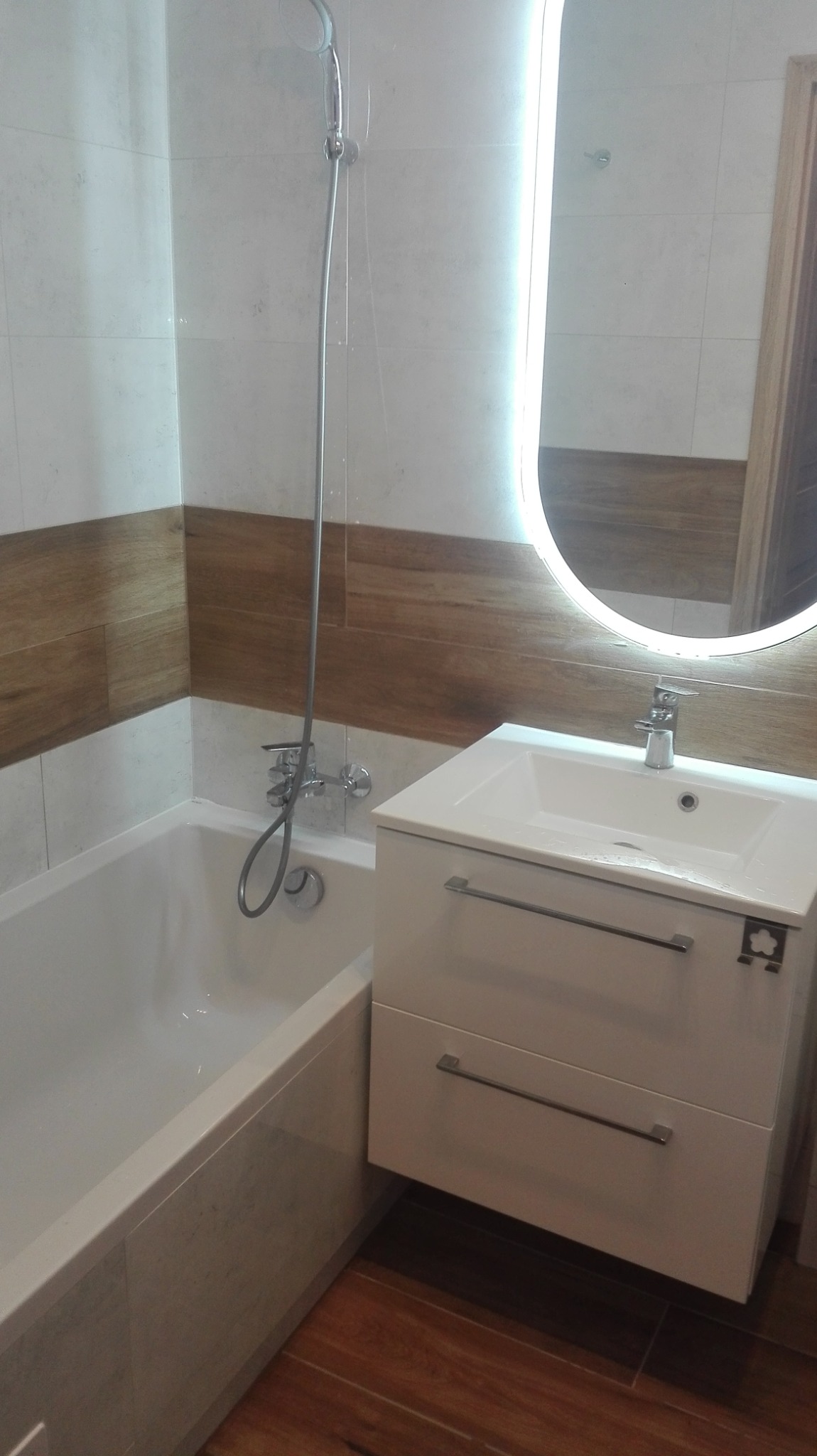 Nowoczesna łazienka z wanną, białą szafką pod umywalką z dwoma szufladami, okrągłym lustrem z podświetleniem LED i mozaiką na ścianach z akcentem drewnopodobnym.