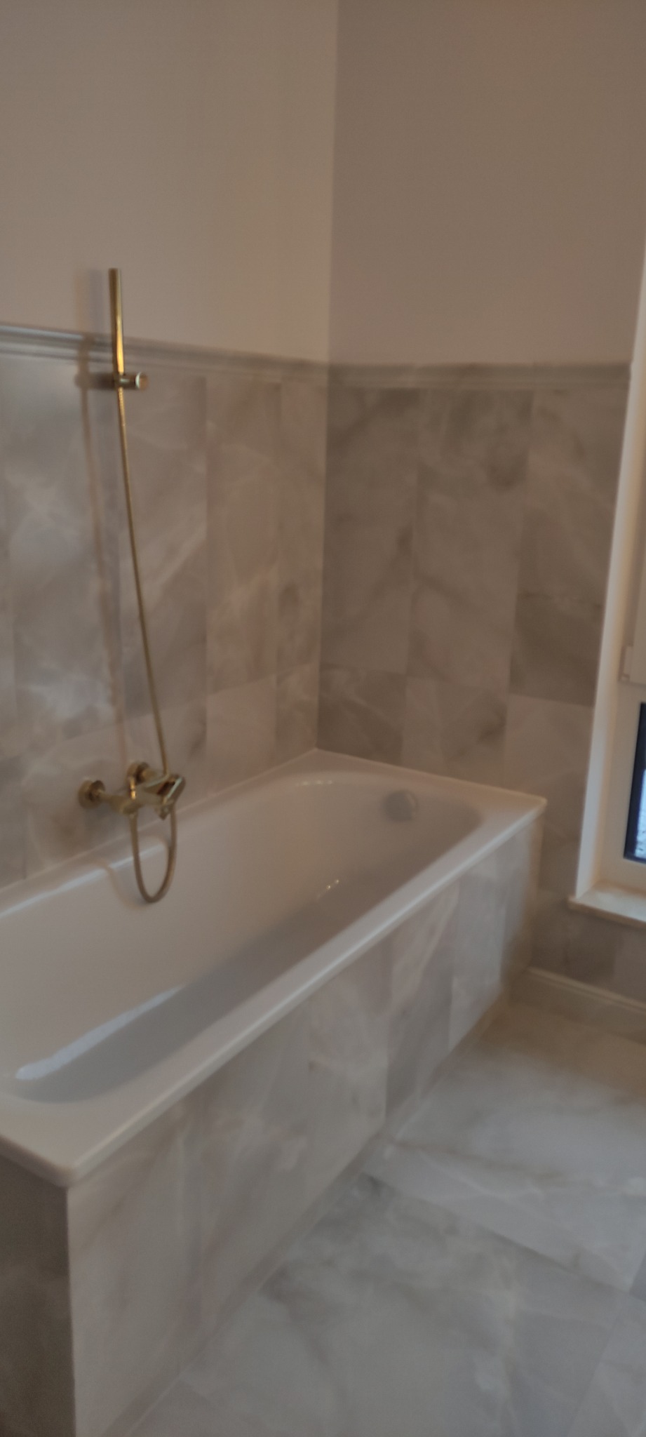 Wykończona łazienka z wanną obudowaną marmurowymi płytkami, złotą baterią prysznicową i marmurową podłogą