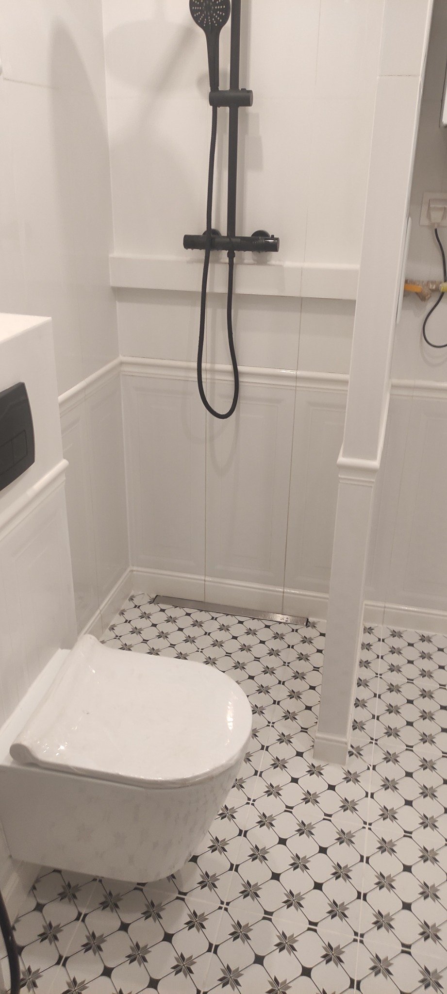 Biała toaleta podwieszana i czarny zestaw prysznicowy w wykończonej łazience z panelami ściennymi i wzorzystą podłogą.