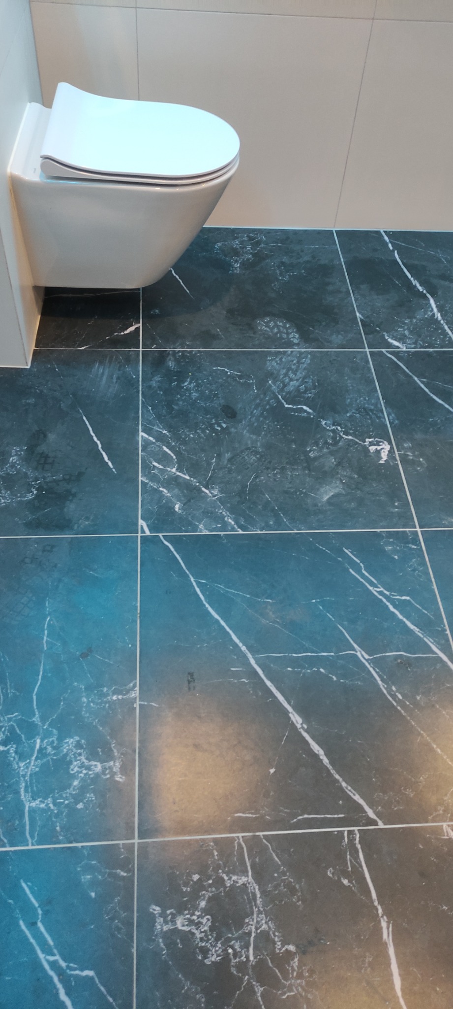 Wisząca toaleta z deską wolnoopadającą i podłoga wyłożona ciemnymi płytkami imitującymi marmur z widocznymi śladami stóp.