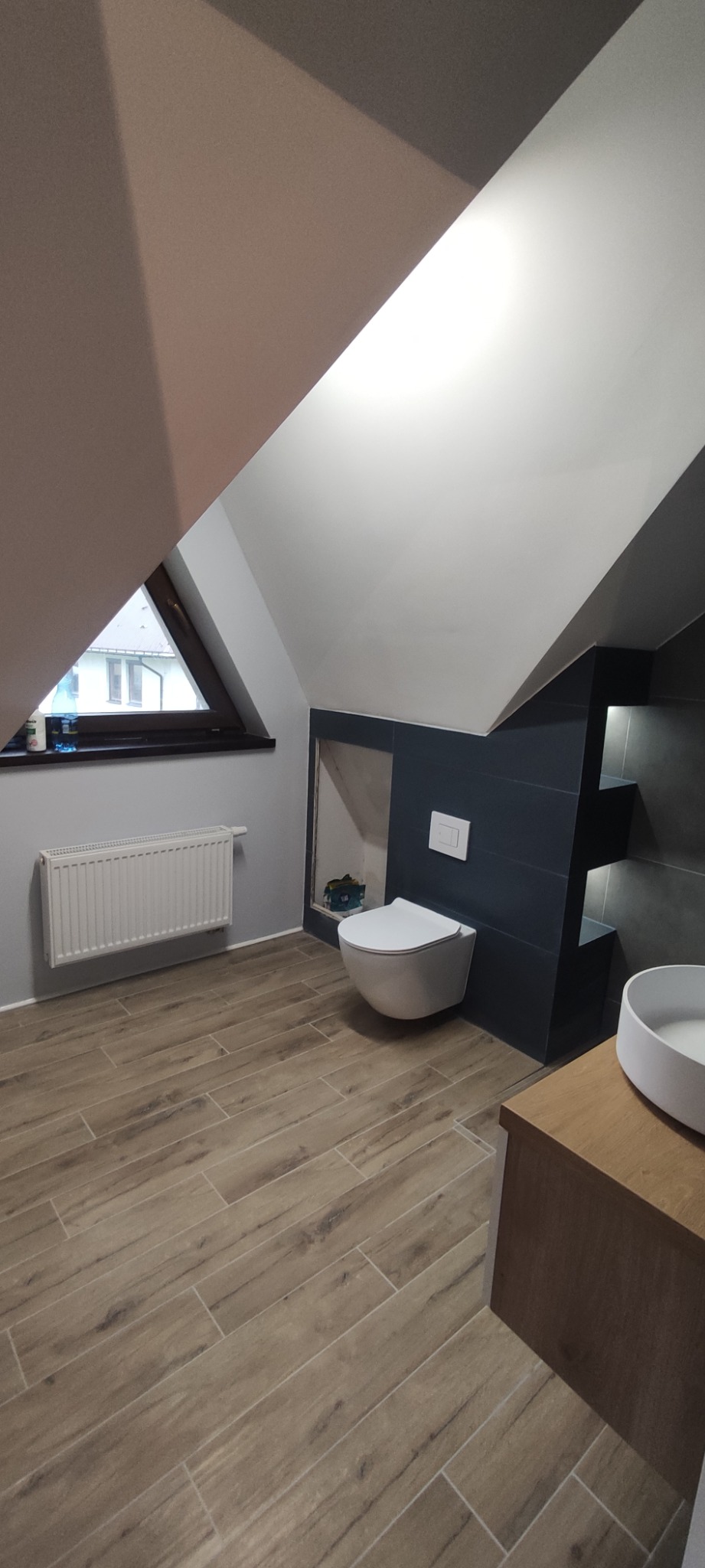 Nowoczesna łazienka na poddaszu z drewnopodobną podłogą, wiszącą toaletą i umywalką nablatową, wykończona ciemnymi płytkami z wnękami.