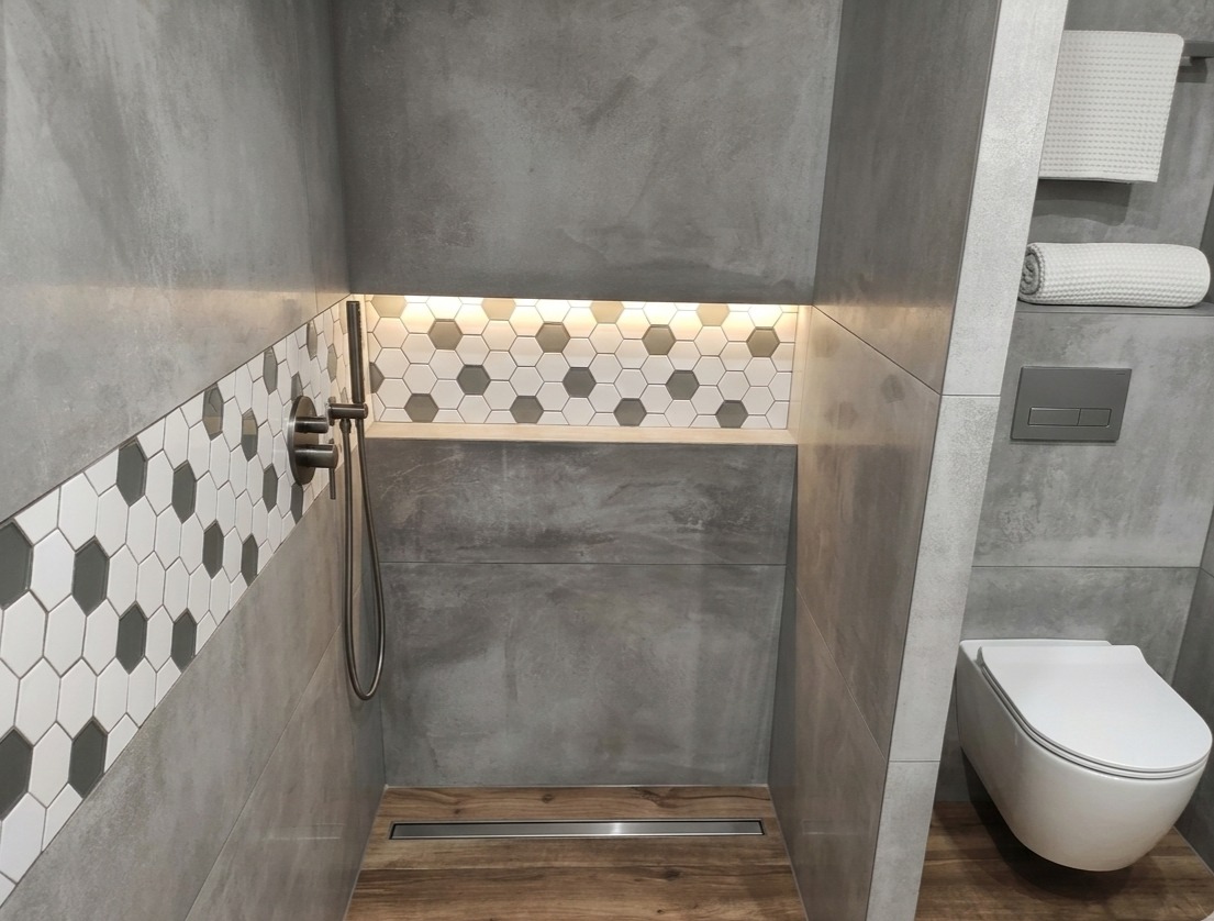 Nowoczesna łazienka z szarą armaturą, heksagonalnymi płytkami i drewnianą podłogą. Kabina prysznicowa typu walk-in z odpływem liniowym. Minimalistyczny design.
