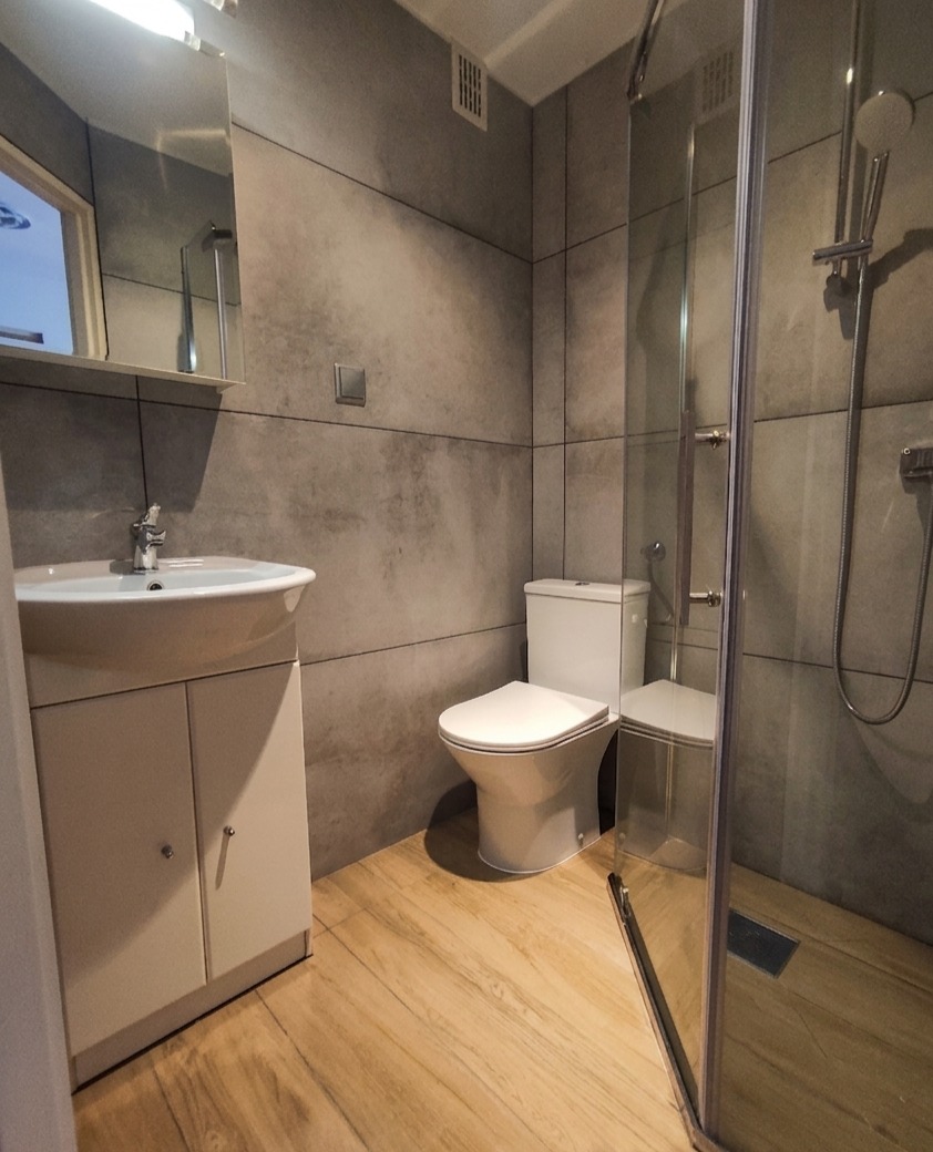 Nowoczesna łazienka z kabiną prysznicową, umywalką z szafką i toaletą. Szare płytki imitujące beton na ścianach, drewnopodobne panele na podłodze. Minimalistyczny design.